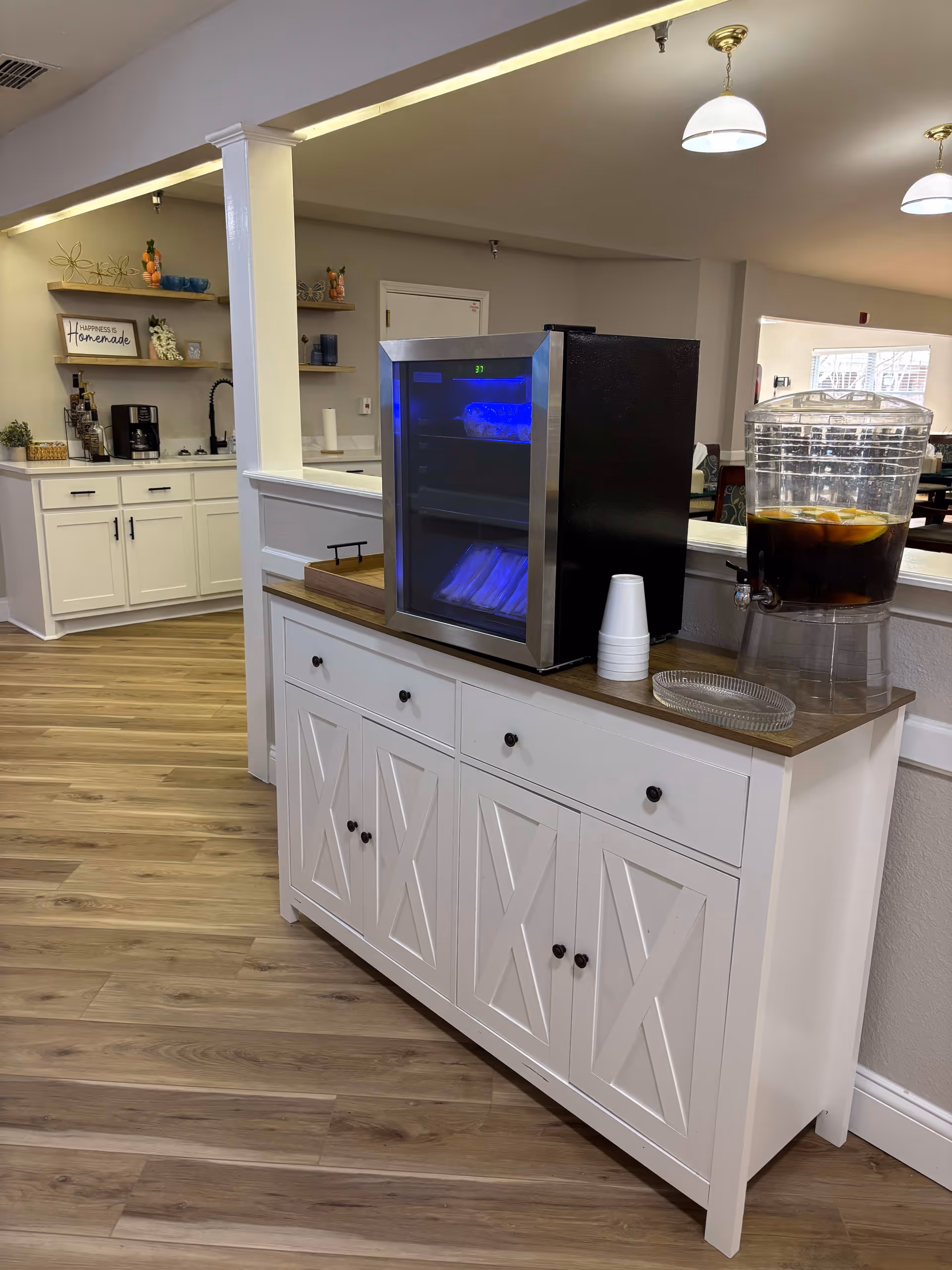 Interior view of a senior living facility area with a white cabinet topped with a small beverage cooler and a large drink dispenser filled with a dark beverage and lemon slices. In the background, there is a kitchen area with white cabinets, shelves holding decorative items, a coffee maker, and a sign that reads 'Happiness is Homemade'. The floor is wood, and the ceiling has hanging light fixtures.