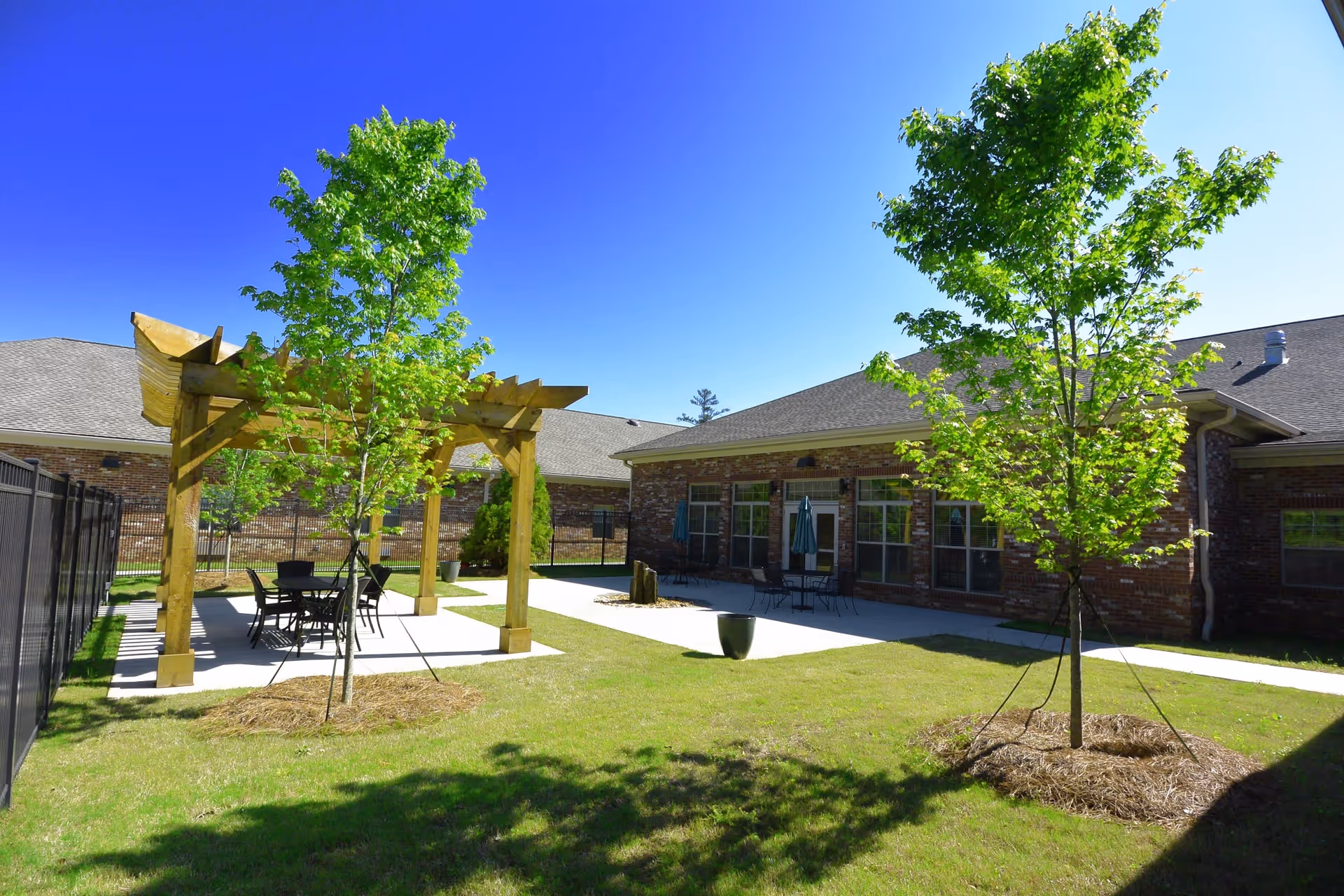 Outdoor courtyard area at Hope Center Memory Care featuring a wooden pergola with a table and chairs underneath, surrounded by green grass and young trees, with a brick building in the background under a clear blue sky.