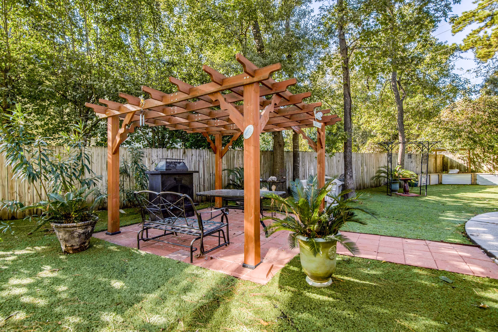 Outdoor garden area with a wooden pergola covering a seating area that includes a metal bench and table. The space is surrounded by green grass, potted plants, trees, and a wooden fence in the background under a clear sky.