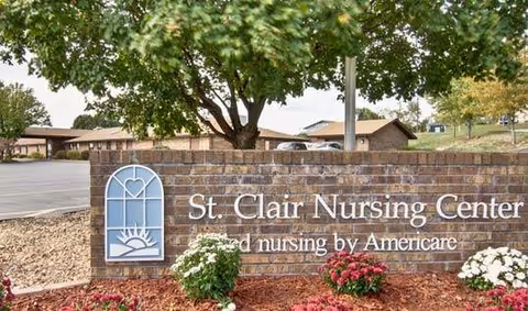 Brick sign for St. Clair Nursing Center with a decorative window and heart logo, surrounded by landscaping with flowers and a large tree in the background.
