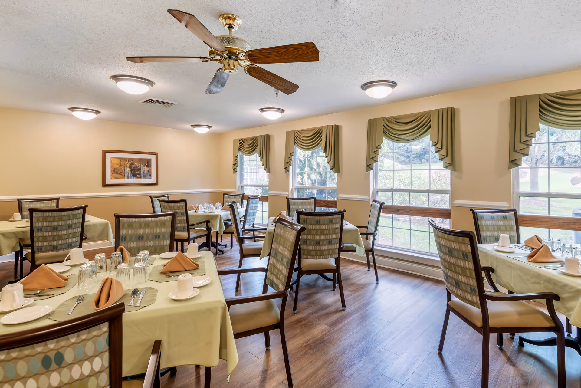 Bright dining room with several tables set for a meal, patterned chairs, green tablecloths, large windows with valances, and a ceiling fan.