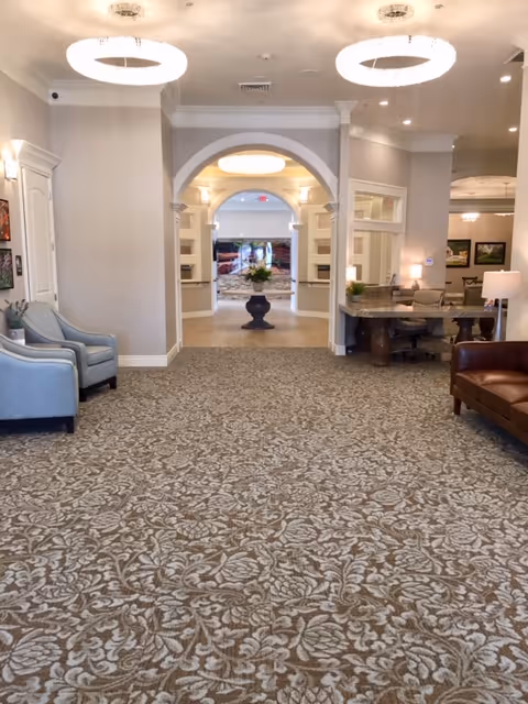 Spacious and well-lit interior common area with patterned carpet flooring, two modern circular ceiling lights, an archway leading to another room with a large decorative vase in the center, light-colored walls, a pair of blue upholstered chairs on the left, a brown leather sofa on the right, and a desk area with chairs and a lamp on the right side.
