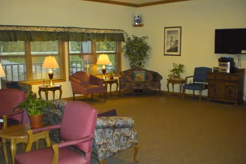 A cozy living room area in Meadow View Assisted Living with several upholstered chairs and sofas arranged around small wooden tables. Two table lamps are lit near a large window with a valance, and there is a potted plant on one of the tables. A framed picture hangs on the wall, and a flat-screen TV is mounted above a wooden cabinet with electronic equipment.
