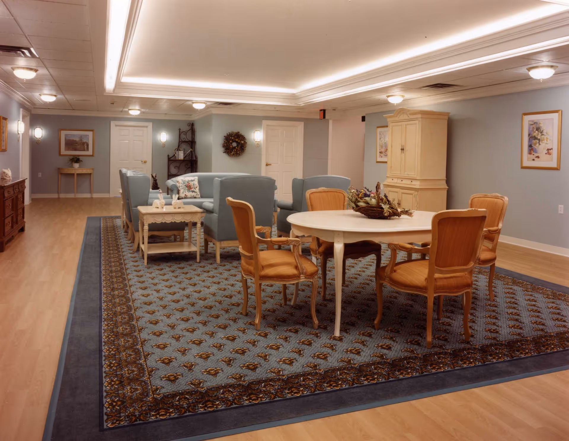 A cozy common area in an assisted living facility featuring a large patterned area rug, a round white table with four wooden chairs, and a seating area with blue upholstered sofas and armchairs. The room has soft lighting, framed artwork on the walls, and light wood flooring.