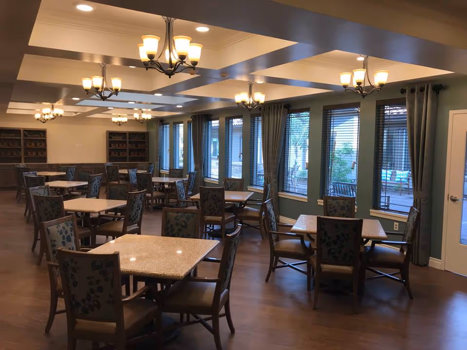Spacious dining room with multiple square tables and patterned chairs, chandeliers overhead, large windows with blinds and curtains, and wood flooring.