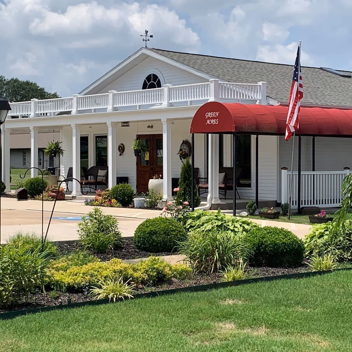 Exterior view of a single-story white building with a red awning labeled 'Green Acres'. The building has a porch with seating, white railings, and an American flag on a pole. There are well-maintained shrubs and plants in the foreground under a partly cloudy sky.