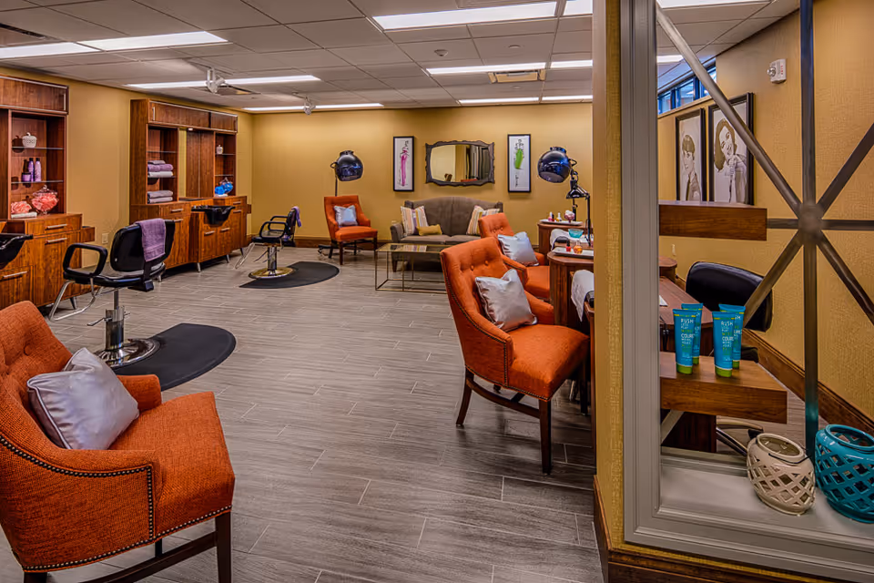 Interior view of a salon area in a senior living facility with orange upholstered chairs, hair styling stations with mirrors and hair dryers, a small seating area with a sofa and framed artwork on the walls, and shelves with towels and hair products.