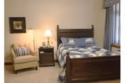 A cozy bedroom with a wooden bed frame, a neatly made bed with a blue and white checkered comforter and pillows, a wooden nightstand with a lamp and decorative items, a beige armchair with striped cushions, and a window with blinds and a valance.