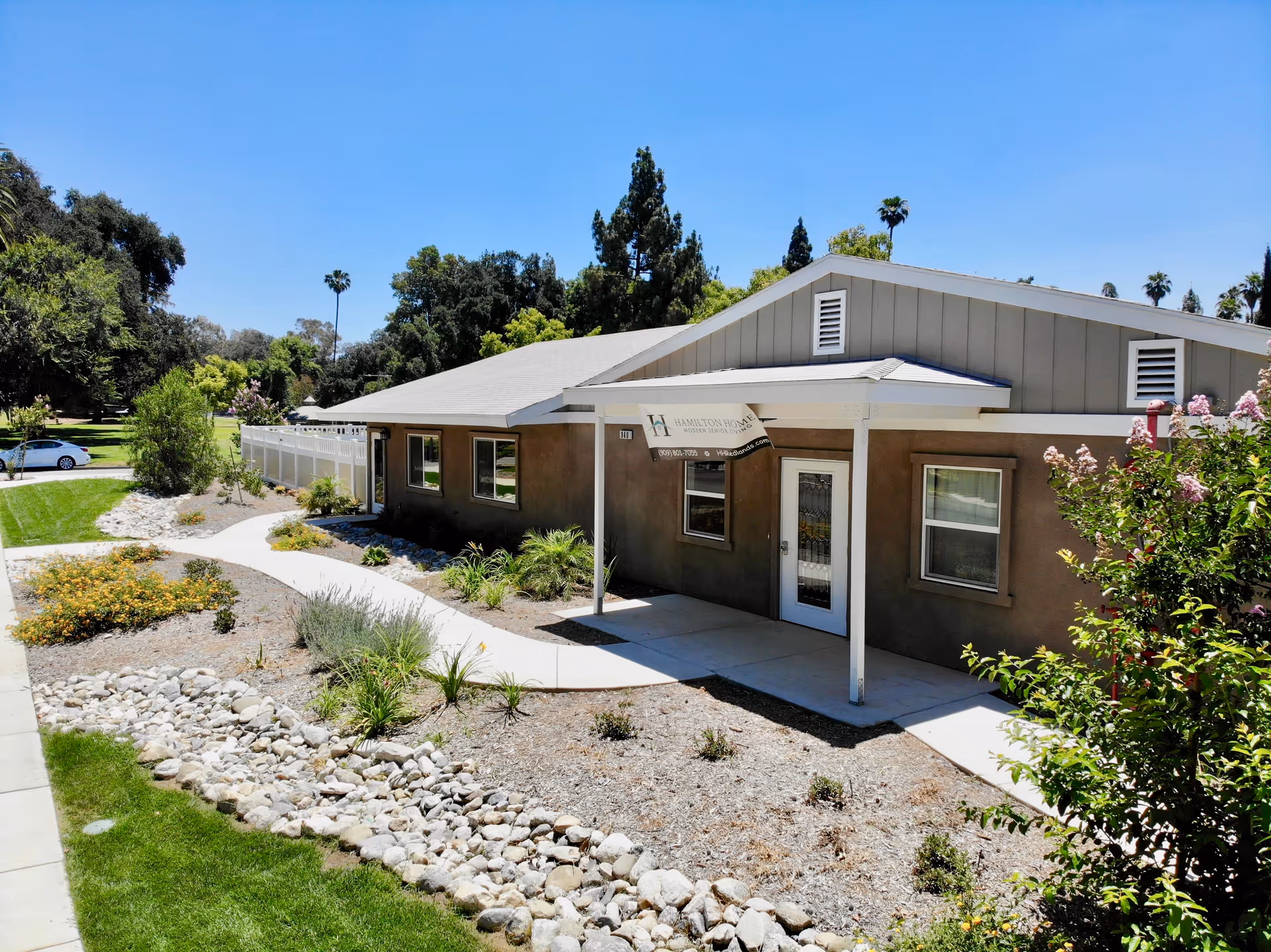 Exterior view of a single-story building with a gray roof and beige walls, surrounded by landscaped garden beds with rocks, plants, and shrubs. A concrete pathway leads to a covered entrance with a small awning. Trees and a clear blue sky are visible in the background.