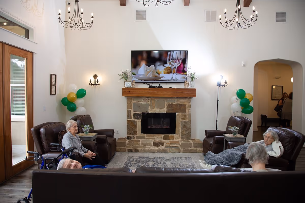 A cozy living room in a senior living facility with four elderly women seated on leather chairs and a sofa around a stone fireplace. A flat-screen TV is mounted above the fireplace, and there are green, white, and gold balloons on either side of the room. The room has high ceilings with chandeliers and wall sconces providing light.