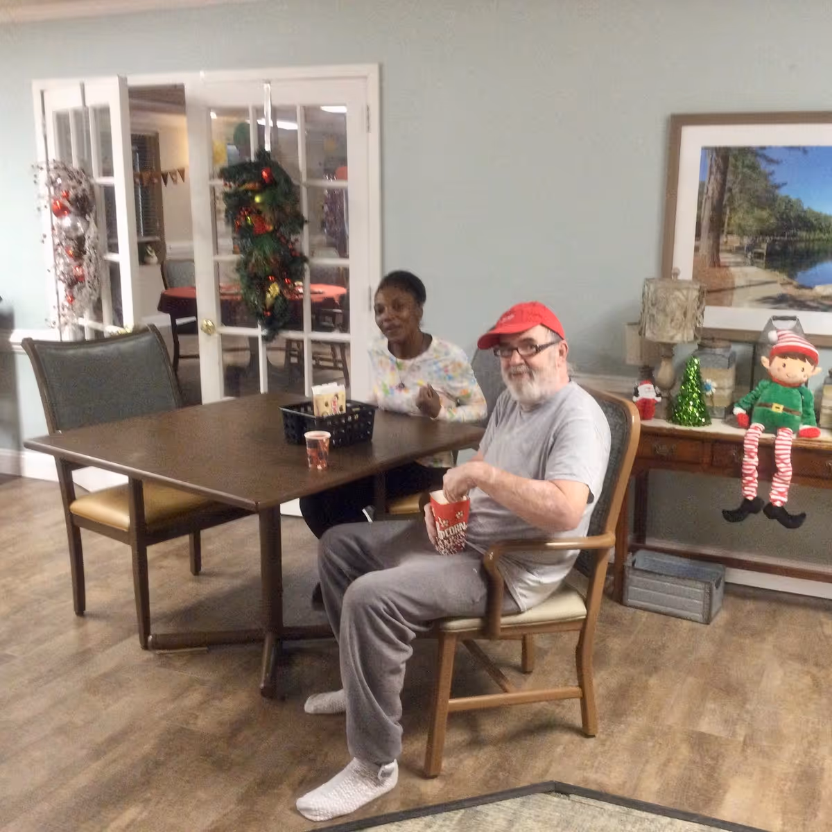 Two people sitting at a wooden table in a room decorated for the holidays. One person is wearing a red cap and holding a popcorn container, while the other is smiling. The room has light-colored walls, a wooden floor, a side table with holiday decorations including a small Christmas tree and an elf doll, and a framed picture on the wall. French doors with holiday wreaths are visible in the background.