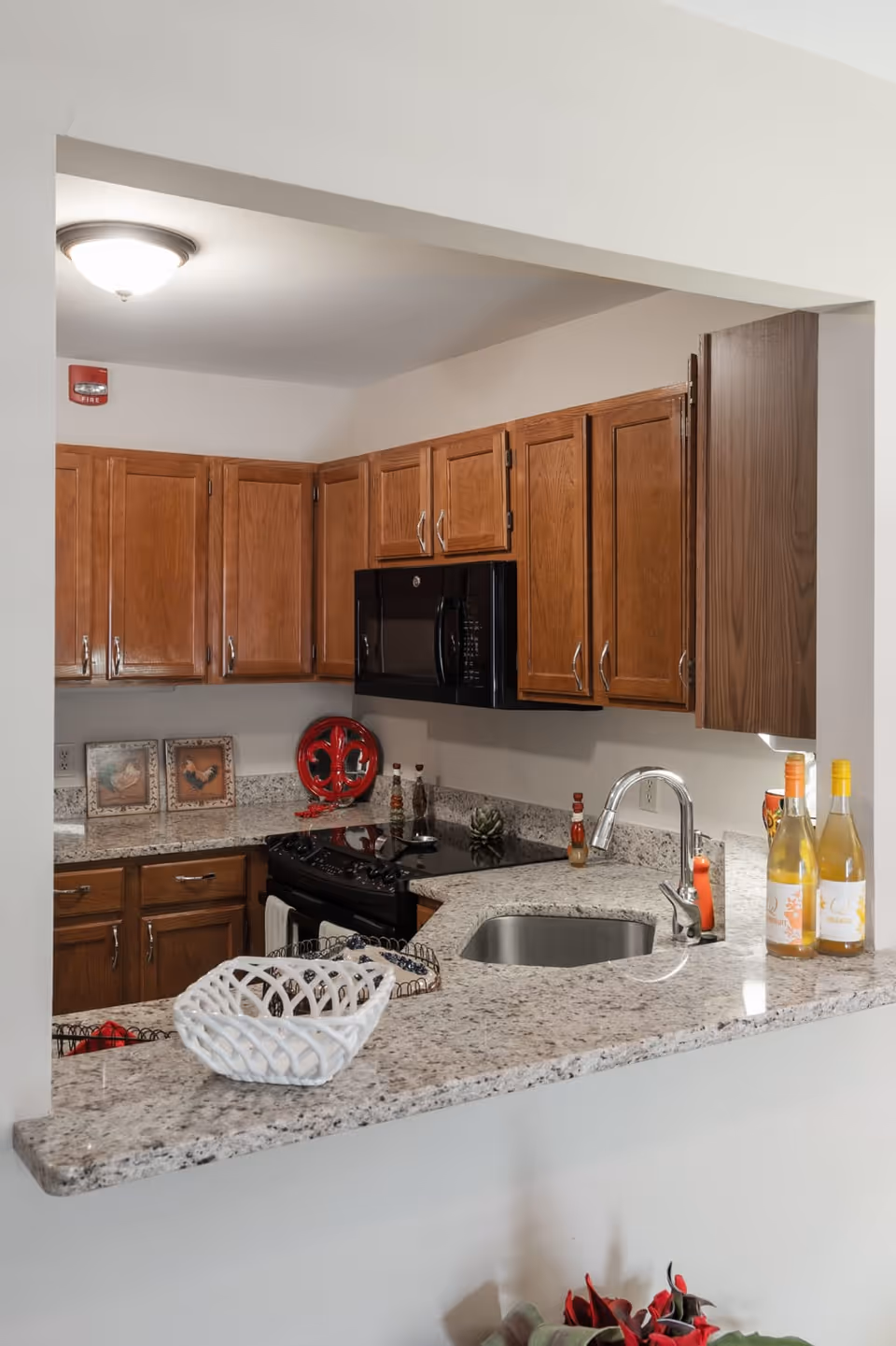 A kitchen with wooden cabinets, a black microwave above a black stove, and a granite countertop with a built-in sink. On the countertop are a white decorative basket, three bottles of wine, and some small decorative items. The kitchen has a light fixture on the ceiling and framed pictures on the counter against the wall.