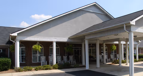 Exterior view of a senior living facility entrance with a covered drop-off area supported by white columns, brick walls, multiple windows, and several hanging plants. Rocking chairs are arranged along the front porch under the covered area.