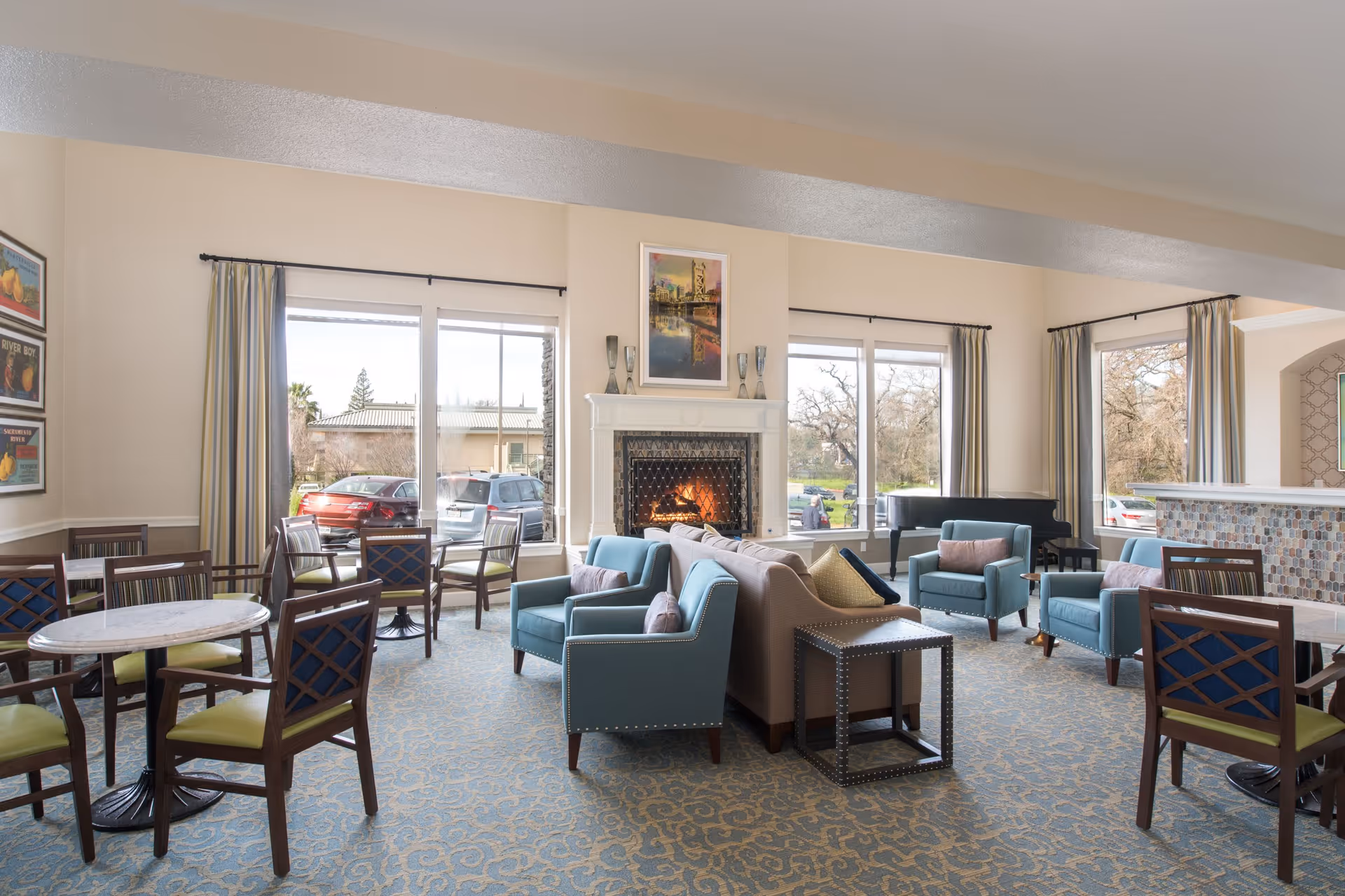 A bright and spacious common area with multiple seating options including blue armchairs, a beige sofa, and wooden chairs around small round tables. There is a lit fireplace in the center with a painting above it, large windows with curtains letting in natural light, and a piano in the corner. The room has a patterned carpet and light-colored walls.