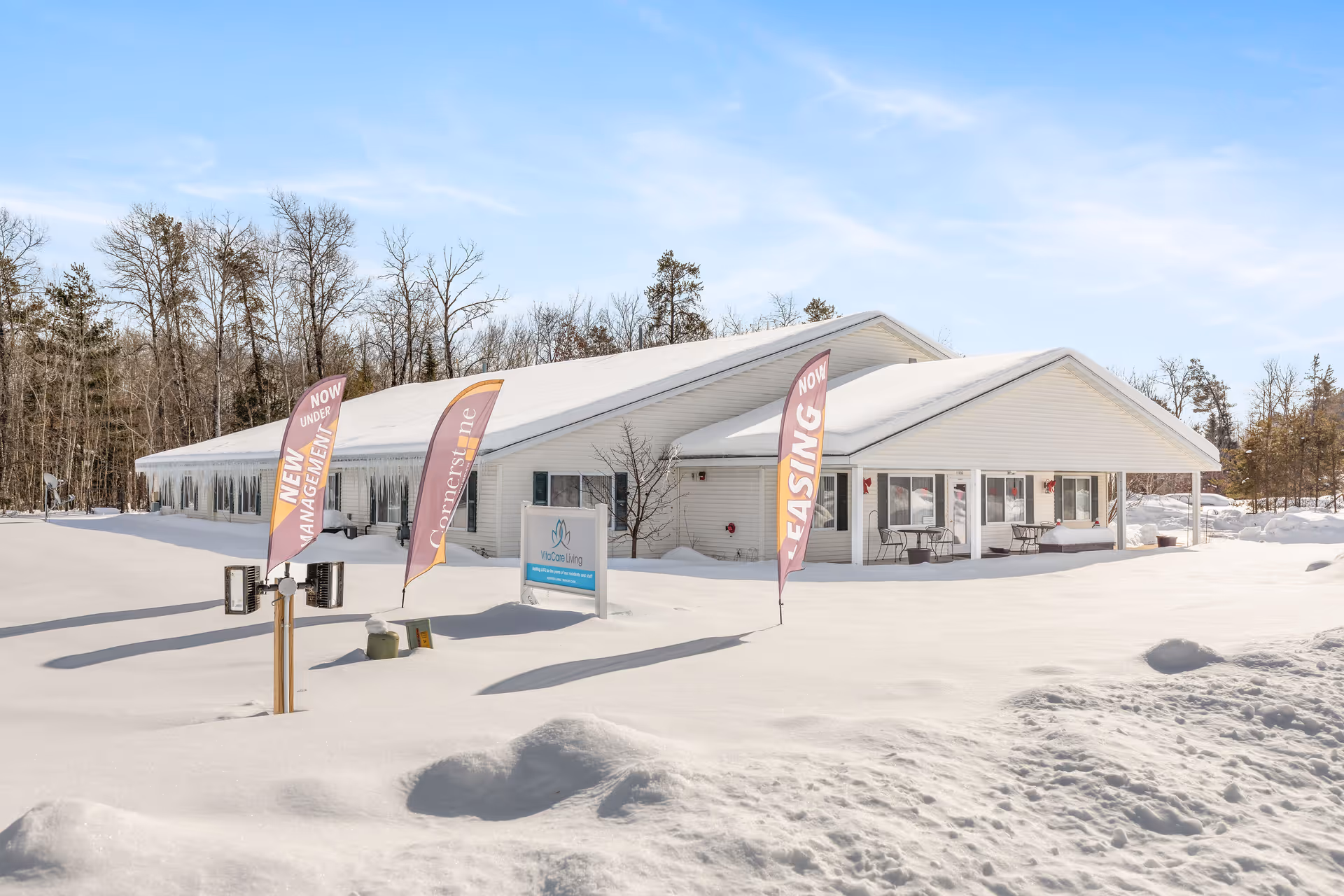 Single-story white senior living building with promotional flags and a sign in front, surrounded by snow.