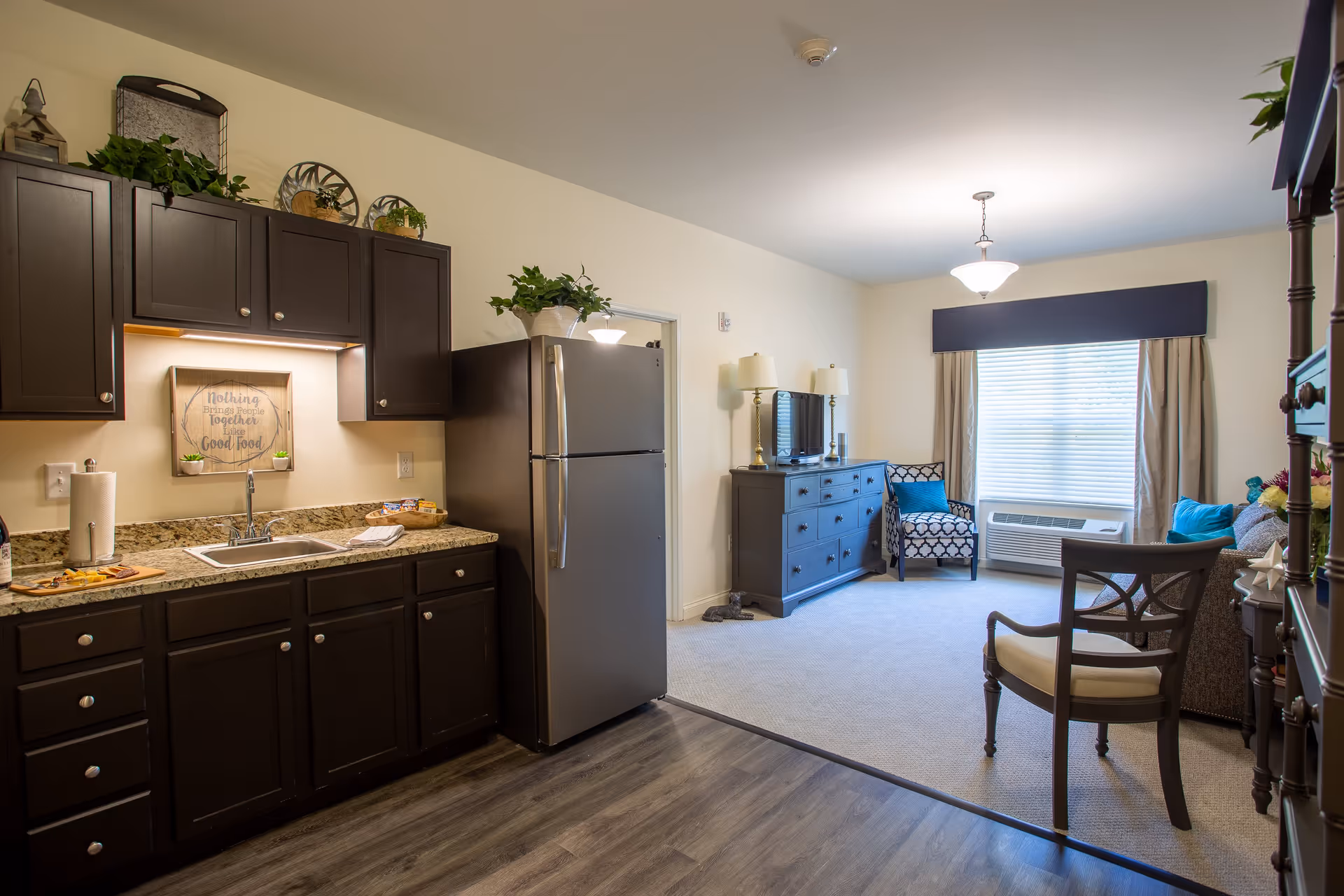 A bright and cozy interior space featuring a kitchen area with dark wood cabinets, a granite countertop, and a stainless steel refrigerator. The kitchen opens into a living room with a carpeted floor, a window with blinds and curtains, a TV on a dark dresser, two table lamps, a patterned armchair with a blue cushion, a sofa with blue pillows, and a wooden chair in the foreground.