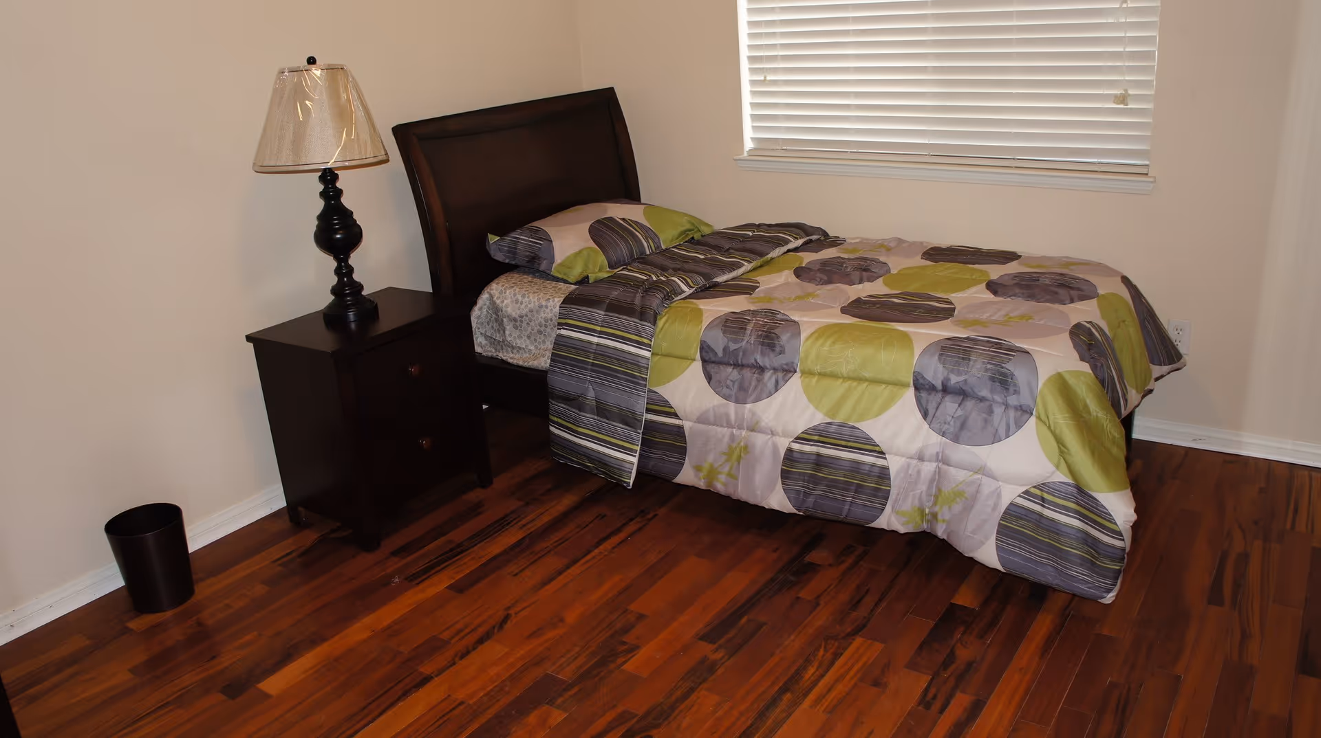 A small bedroom with a single bed covered in a comforter with green, gray, and purple circular patterns. Next to the bed is a dark wooden nightstand with two drawers and a lamp with a beige shade. The room has wooden flooring and a window with closed white blinds.