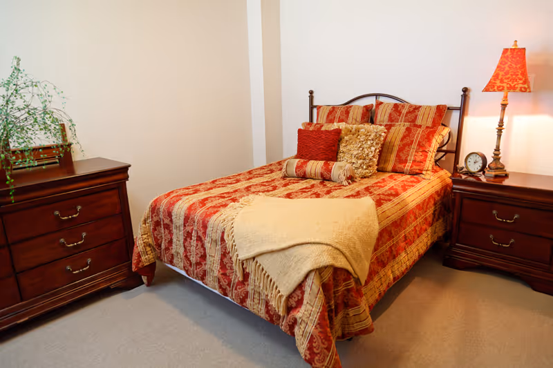 A neatly made bed with red and gold patterned bedding flanked by wooden nightstands, one holding a lamp and clock.