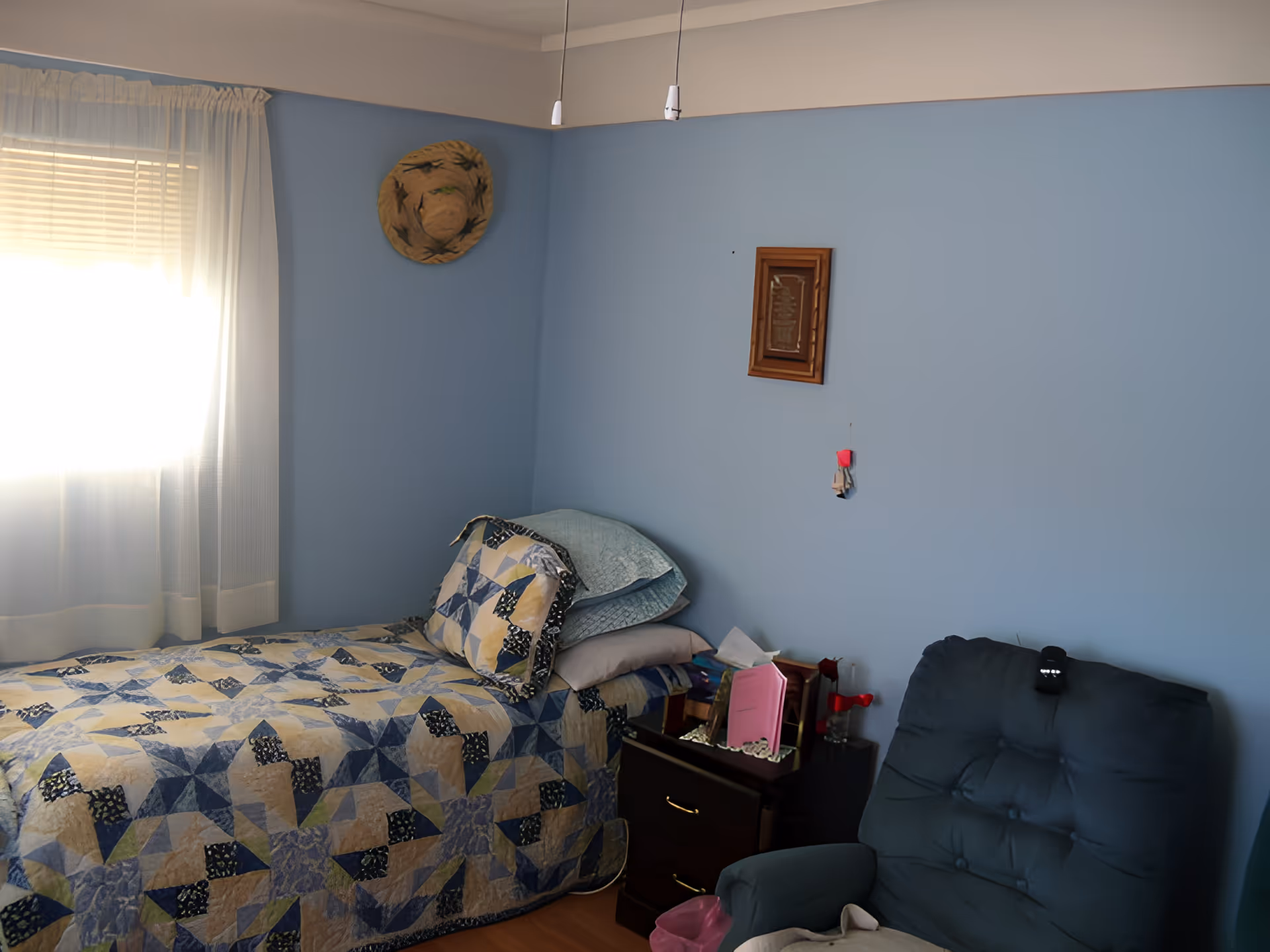 A small bedroom with light blue walls, a single bed with a patterned quilt and pillows, a dark wooden nightstand with various items on it, a blue cushioned armchair, a window with white curtains letting in sunlight, a straw hat hanging on the wall, and a small framed decoration.