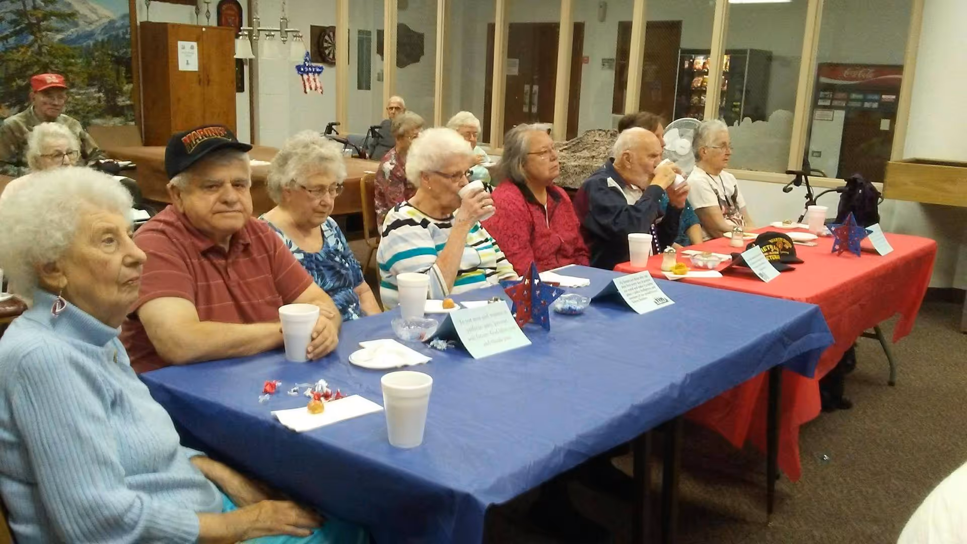 A group of elderly people sitting at tables covered with red and blue tablecloths in a community room. They appear to be attending an event or gathering, with some drinking from white cups and small decorations on the tables. The room has a casual, social atmosphere with various items and furniture in the background.