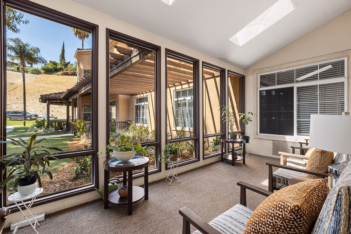 Sunlit enclosed sitting area with large floor-to-ceiling windows, potted plants, and armchairs overlooking a courtyard.