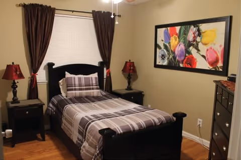 Single bedroom with a dark wood twin bed dressed in gray plaid bedding, matching nightstands and lamps, a dresser, and colorful floral artwork on the wall.
