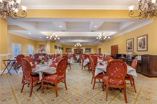 A spacious dining room with multiple round tables covered with white tablecloths and set with napkins and tableware. The chairs are wooden with red patterned upholstery. The room has yellow walls, decorative chandeliers hanging from the ceiling, framed artwork on the walls, and large windows letting in natural light.