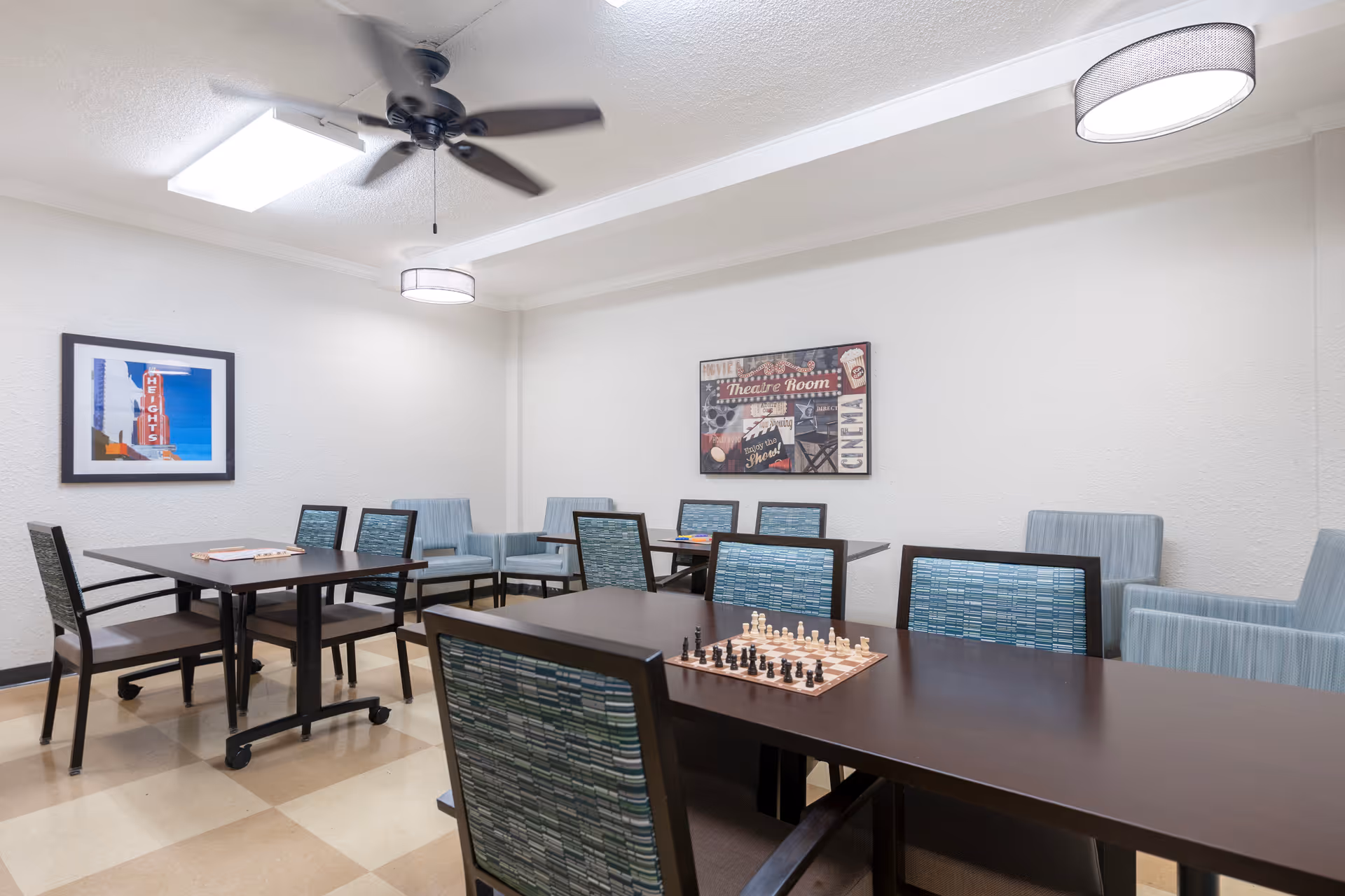 A well-lit room with several tables and chairs arranged for group activities. One table has a chessboard set up with chess pieces in play. The walls are decorated with framed pictures, including one with a theater theme. The room has a ceiling fan and a modern light fixture, with a checkered floor pattern.