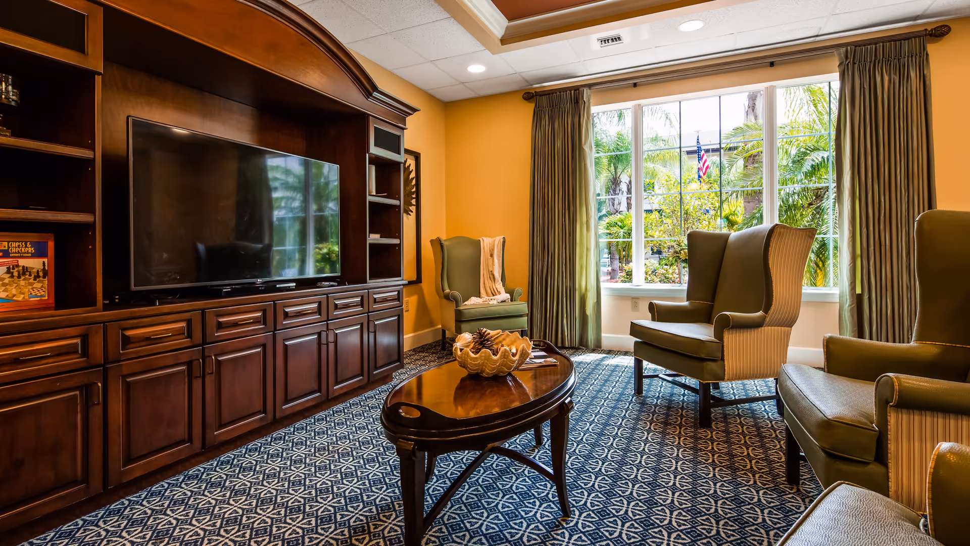 A cozy living room area with a large wooden entertainment center holding a flat-screen TV, three green upholstered armchairs, a wooden coffee table with a decorative bowl, and a large window with green curtains showing a garden with palm trees and an American flag outside.