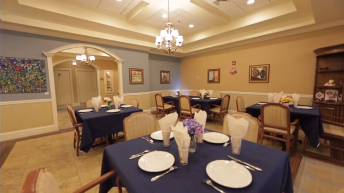 A dining room with several tables covered in dark blue tablecloths, each set with white plates, silverware, and folded napkins in glasses. The room has beige and blue walls adorned with framed artwork, a chandelier hanging from a tray ceiling, and a wooden cabinet with decorative items on the right side.