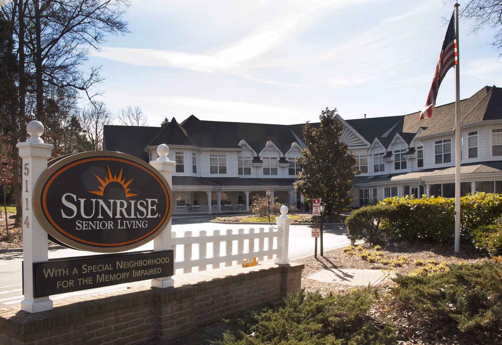 Front exterior of the Sunrise Senior Living facility with a large entrance sign, white fence, and American flag.