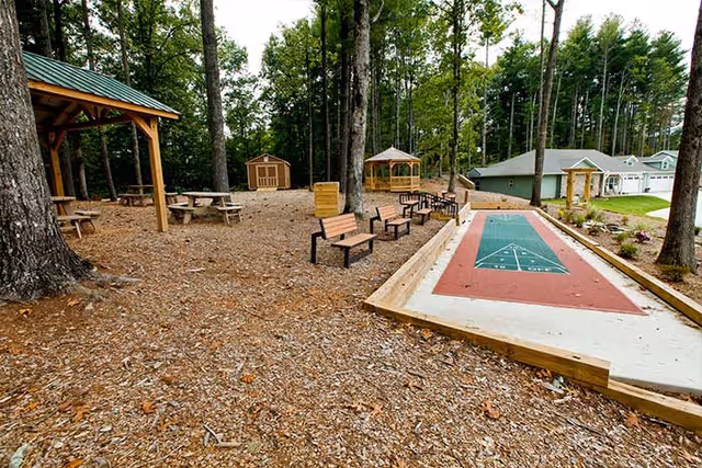 Outdoor recreational area with a shuffleboard court, several benches, picnic tables under a wooden pavilion, a gazebo, and a small shed surrounded by tall trees and residential buildings in the background.