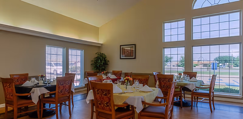 Bright dining room with several set tables and wooden chairs beside large windows overlooking the outside.