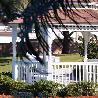 White gazebo with a brown shingled roof surrounded by green grass, bushes, and trees in an outdoor garden area.
