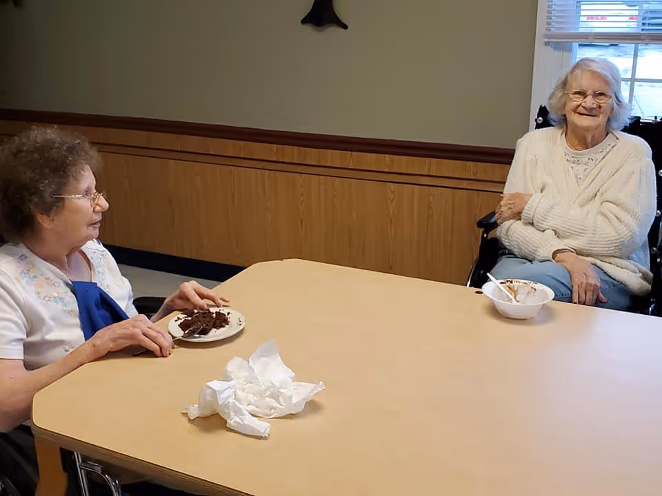 Two elderly women sitting at a wooden table in a room with wood paneling and a window. One woman is eating chocolate cake from a plate, and the other has a bowl with a spoon. Both women are smiling and appear to be enjoying their time together.