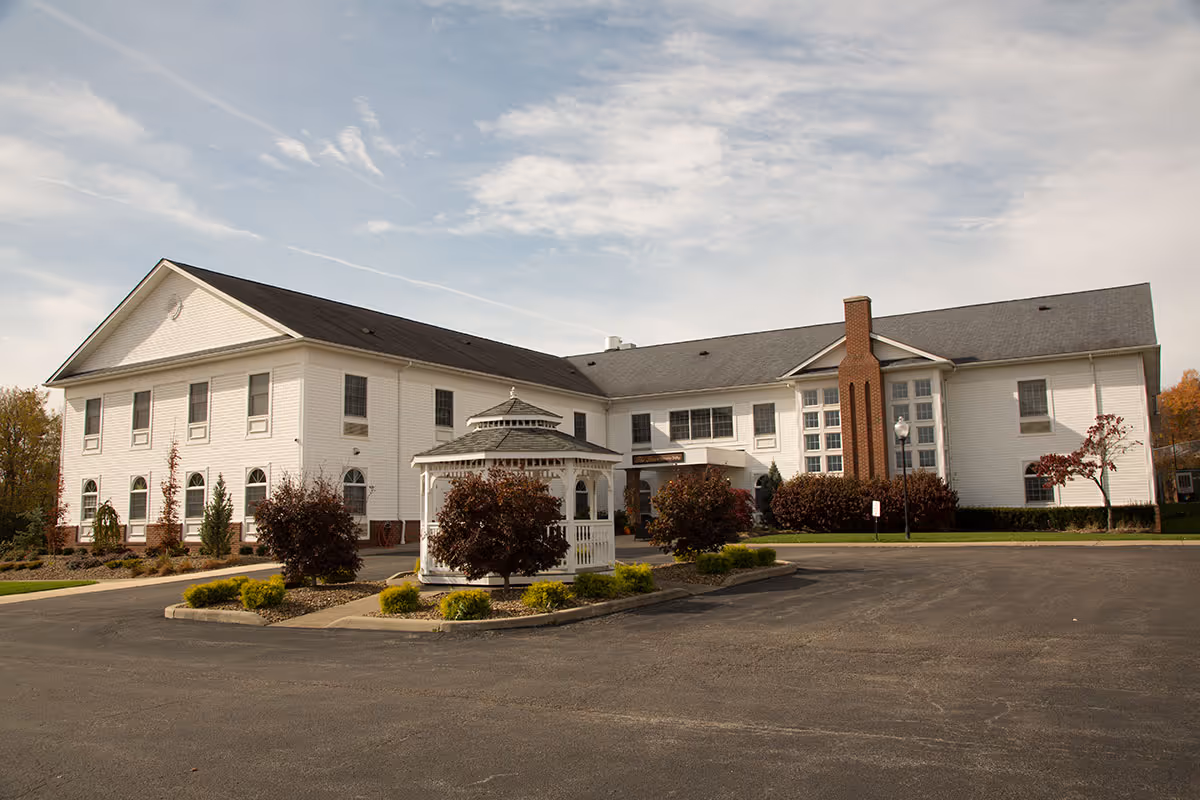 Exterior view of a large two-story white building with a gray roof, surrounded by landscaped bushes and small trees. There is a white gazebo in front of the building and a paved driveway or parking area in the foreground under a partly cloudy sky.