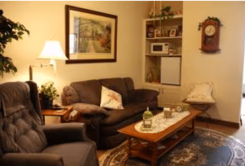 A cozy living room with a brown sofa and a matching armchair. A wooden coffee table with decorative items sits on a patterned rug. A floor lamp and a framed picture are on the wall behind the sofa. A built-in shelving unit holds a microwave, mini fridge, and various decorative items. A wall clock is mounted on the adjacent wall.