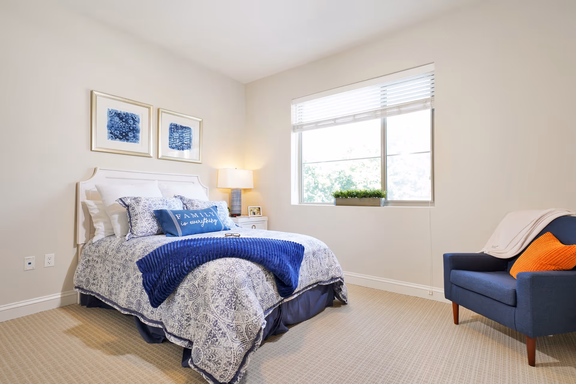 A bright and clean bedroom with a neatly made bed featuring blue and white patterned bedding and a decorative pillow that says 'Family is everything.' There are two framed blue artworks above the bed, a bedside table with a lamp and a small photo frame, a large window with blinds letting in natural light, and a blue armchair with an orange pillow and a light-colored throw blanket.