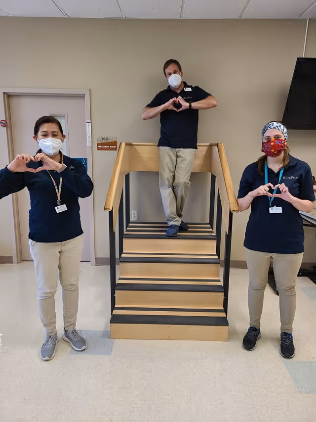 Three staff members wearing masks stand indoors in front of a small wooden staircase with black railings. Two staff members stand on either side of the staircase making heart shapes with their hands, while the third stands on the stairs also making a heart shape with his hands. They are dressed in navy blue tops and beige pants. A door labeled 'Treatment Room' is visible in the background.