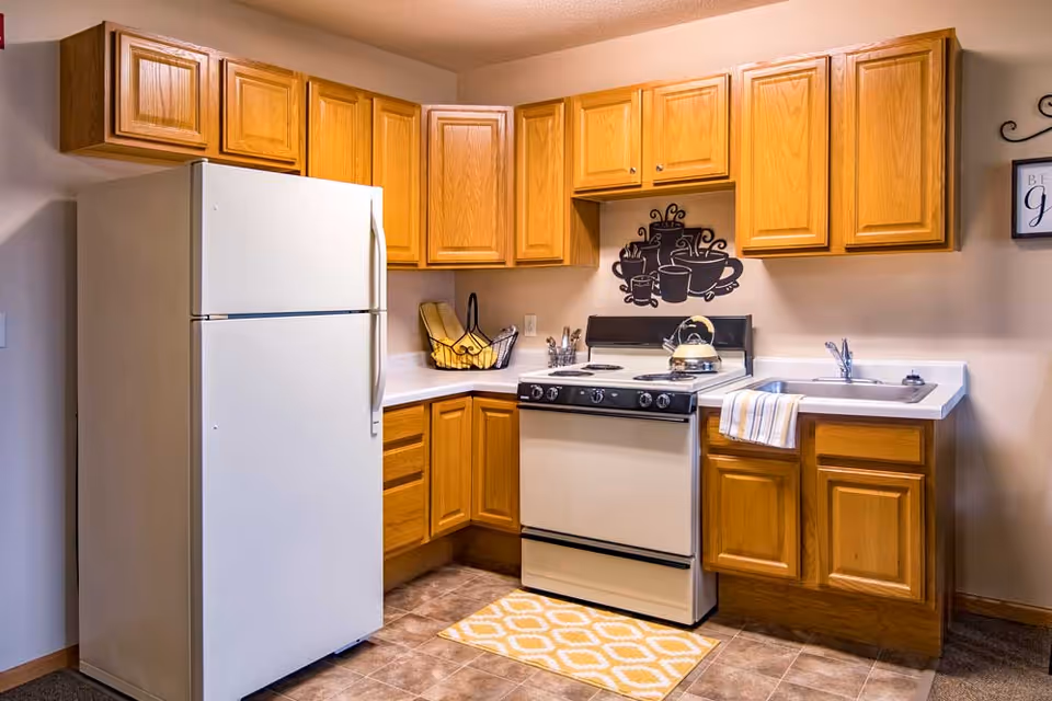 Compact kitchen with oak cabinets, a white refrigerator, a stove with kettle, sink, and a decorative coffee-themed wall decal.