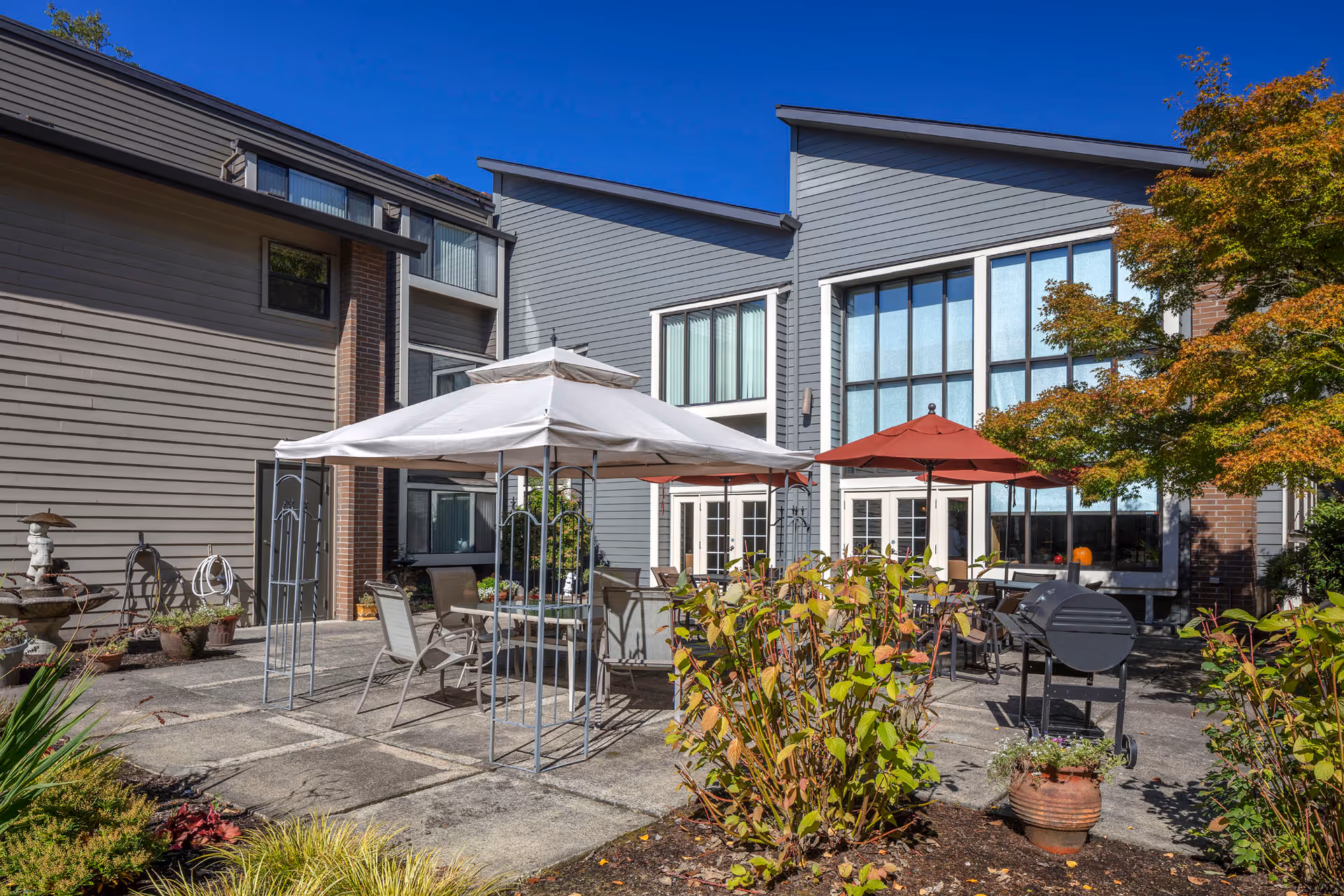 Outdoor patio area at Creekside Village Retirement Residence featuring a white canopy over a table with chairs, a red umbrella shading another seating area, a barbecue grill, potted plants, and a tree with autumn-colored leaves, all set against the backdrop of a modern building with large windows under a clear blue sky.