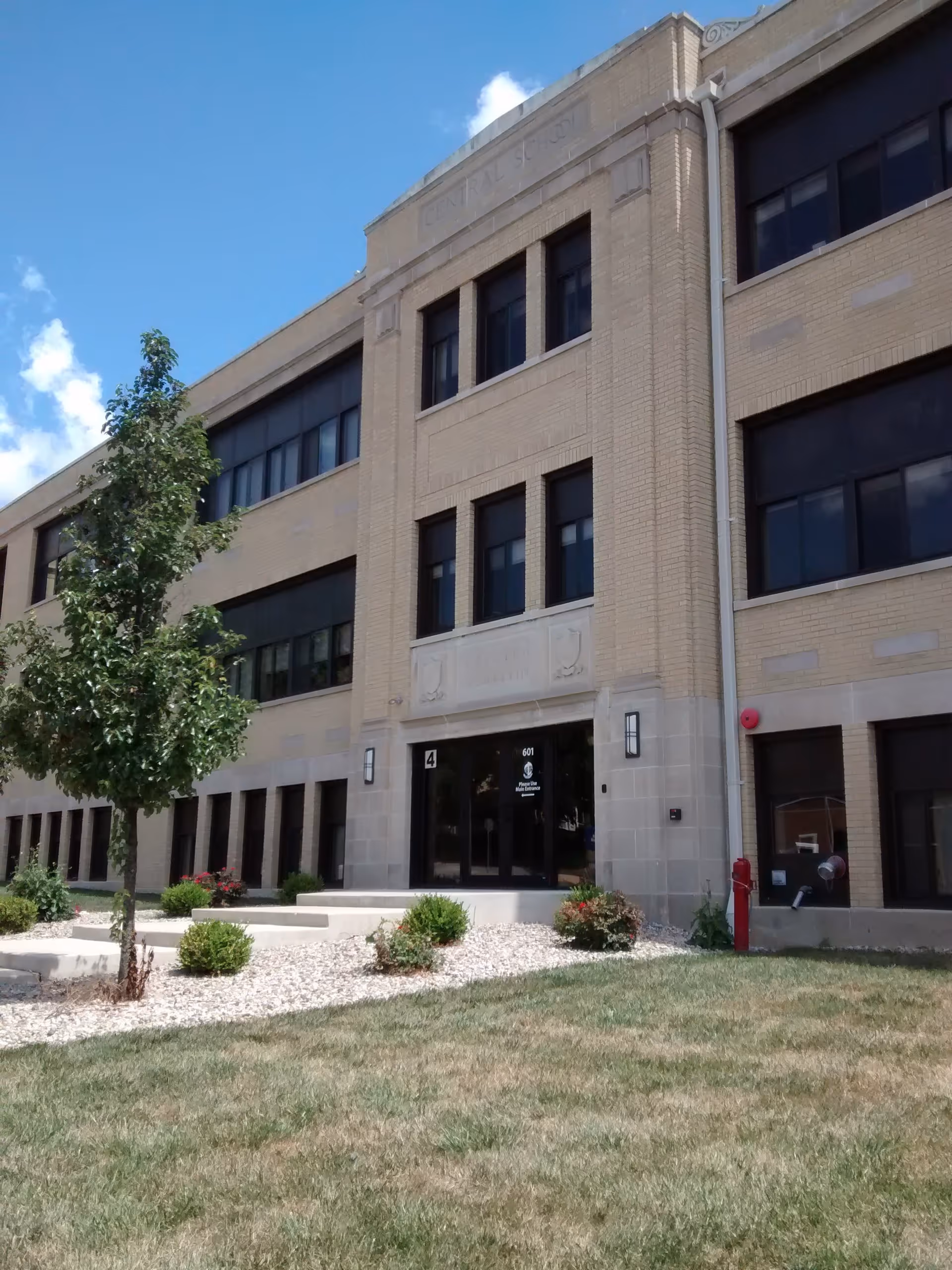 Exterior view of a three-story beige brick building with multiple windows and a main entrance with glass doors. There is a small landscaped area with bushes, a tree, and a grassy lawn in front of the building under a partly cloudy blue sky.