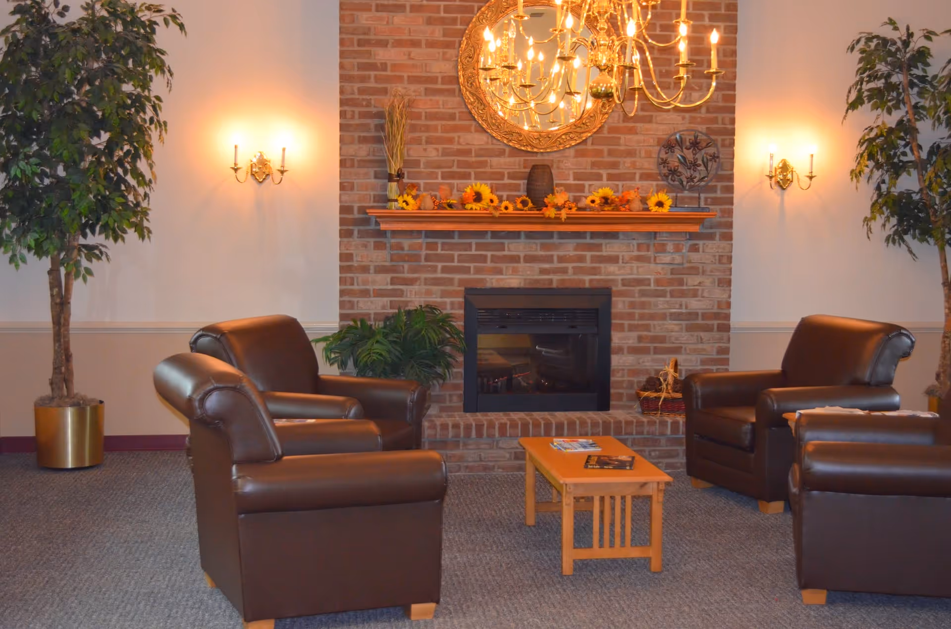A cozy living room area with four brown leather armchairs arranged around a small wooden coffee table. The room features a brick fireplace decorated with sunflowers and a round ornate mirror above it. Two wall sconces with lit candles flank the fireplace, and a chandelier hangs from the ceiling. There are two large potted plants on either side of the fireplace.