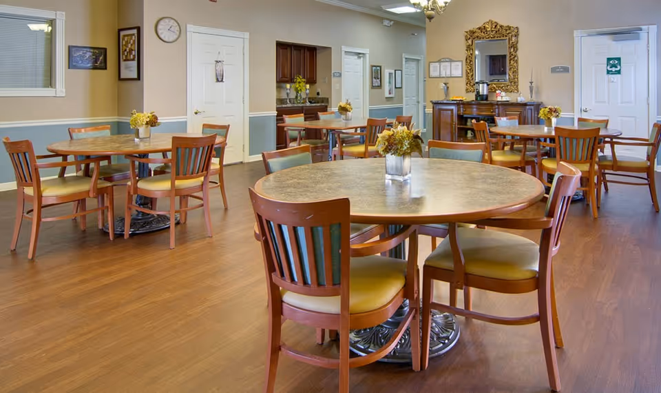 A dining room with several round tables and wooden chairs with yellow cushions. Each table has a small floral centerpiece. The room has wood flooring, beige walls with white trim, and several doors and framed pictures on the walls. A sideboard with a large ornate mirror and coffee station is visible in the background.