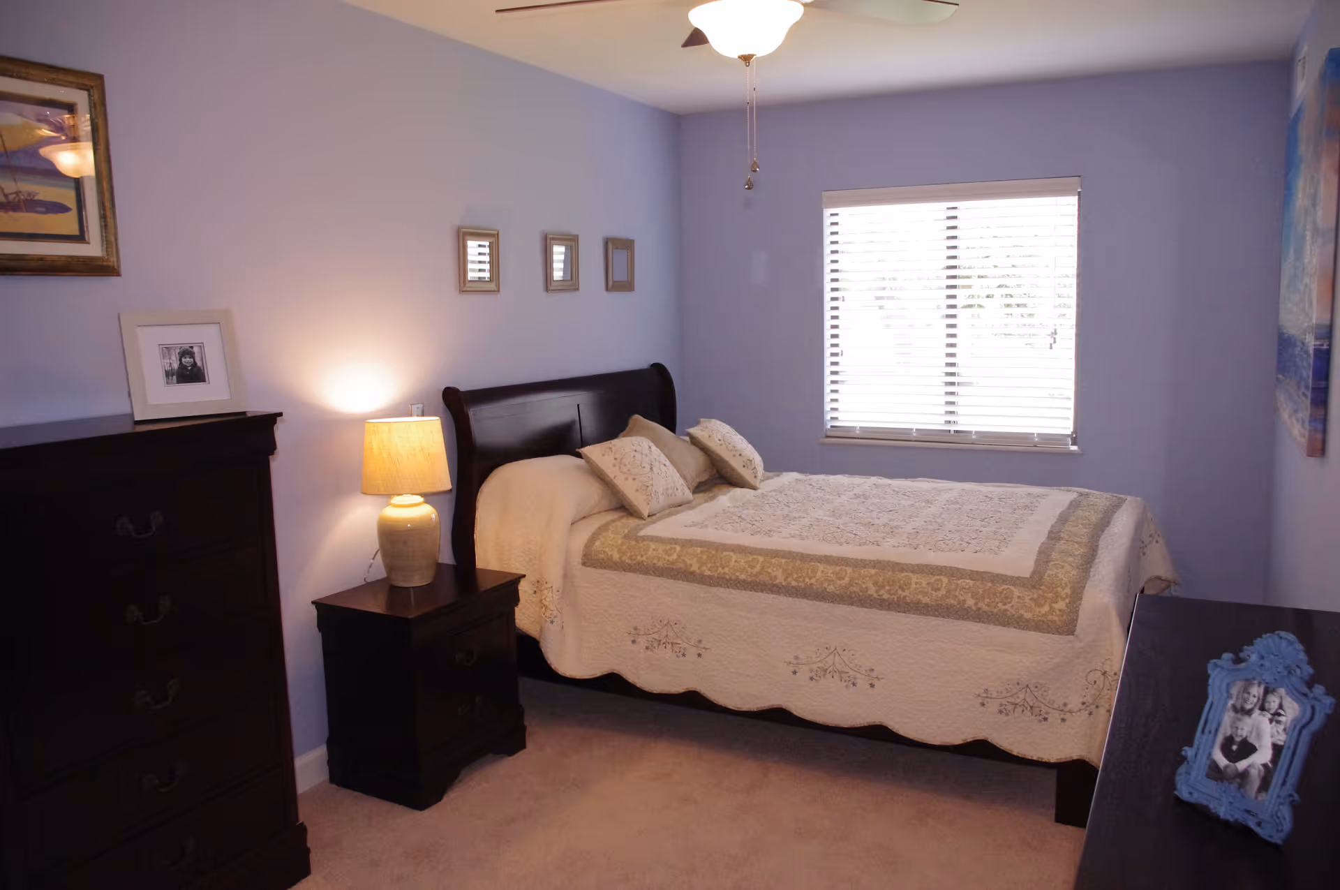 A cozy bedroom with light purple walls, a window with blinds, a bed with a beige and green quilt and two pillows, a wooden nightstand with a lamp, a tall wooden dresser with framed photos on top, and a ceiling fan with a light.