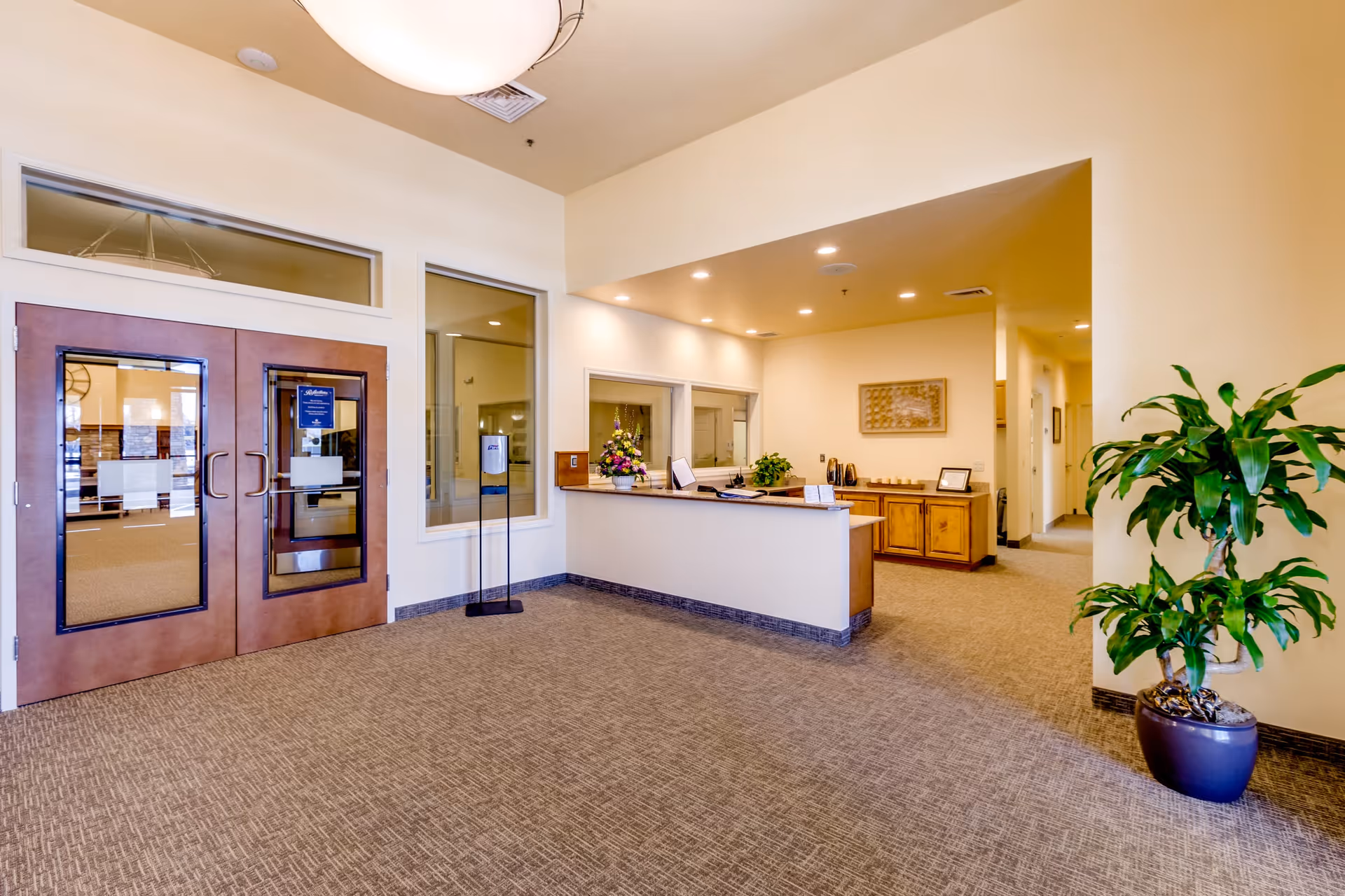 Spacious reception lobby with double glass doors on the left, a front desk in the center-right, and a potted plant on the right.