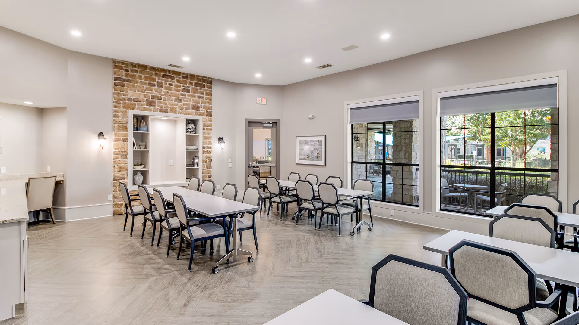 Bright communal dining/activity room with multiple tables and chairs, large windows, and a stone-accent wall.