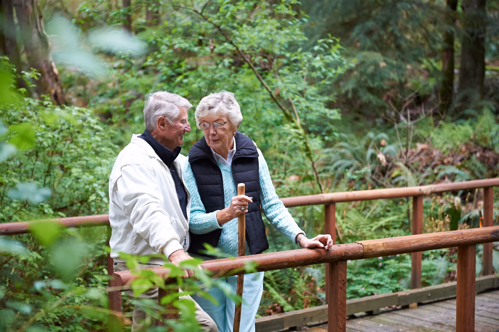 An elderly couple walking together on a wooden bridge surrounded by lush green forest vegetation. The woman is holding a walking stick and wearing a black vest over a light blue sweater, while the man is wearing a white jacket. They appear to be engaged in conversation.