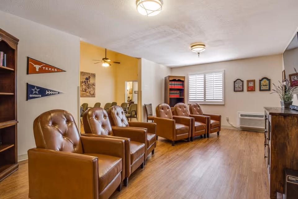 A bright common room with five brown leather armchairs in a row, wooden flooring, bookshelves, wall pennants, and a window with shutters.