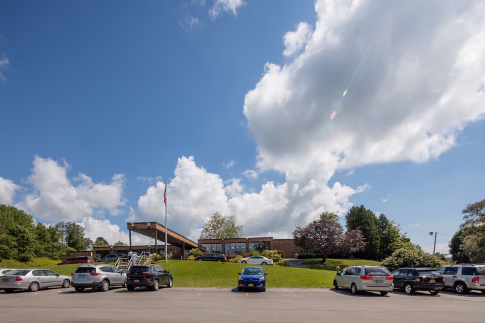 Low brick nursing facility front with a flagpole, grassy lawn and parked cars under a partly cloudy sky.